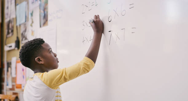 Education, classroom and learning while black student solve a maths equation and write answers on the whiteboard. Smart little school boy doing multiply sum and calculating a solution in class stock photo