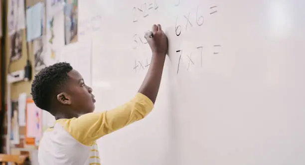 Education, classroom and learning while black student solve a maths equation and write answers on the whiteboard. Smart little school boy doing multiply sum and calculating a solution in class