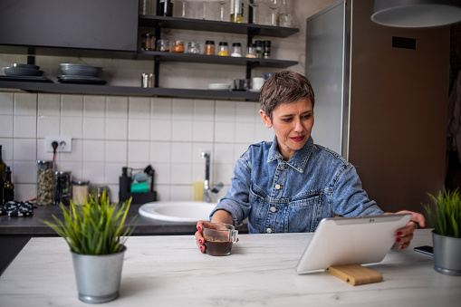 Short-haired mature woman sitting at the bar in her apartment drinking morning coffee and surfing the internet on her tablet