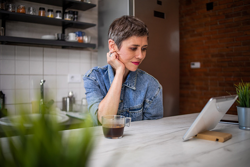 Short-haired mature woman sitting at the bar in her apartment drinking morning coffee and surfing the internet on her tablet