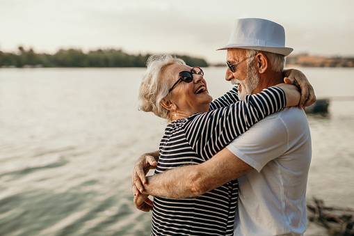 Spontaneous image of a senior couple, enjoying their sunset beach walk together. A cool looking grandpa, wearing a hat, is hugging his beautiful wife, both wearing sunglasses on, smiling and facing each other, feeling romantic. Enjoying conversations, love, and small dance