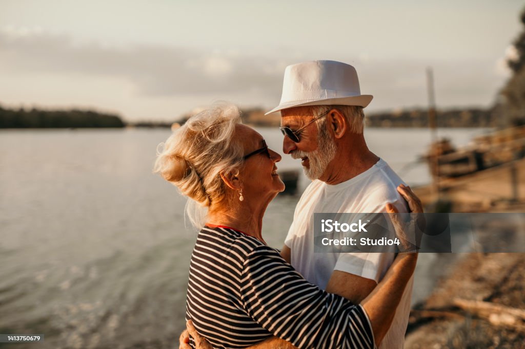 Dancing on the beach on the sunset Spontaneous image of a senior couple, enjoying their sunset beach walk together. A cool looking grandpa, wearing a hat, is hugging his beautiful wife, both wearing sunglasses on, smiling and facing each other, feeling romantic, dancing. Enjoying conversations, love, and small dance 65-69 Years Stock Photo