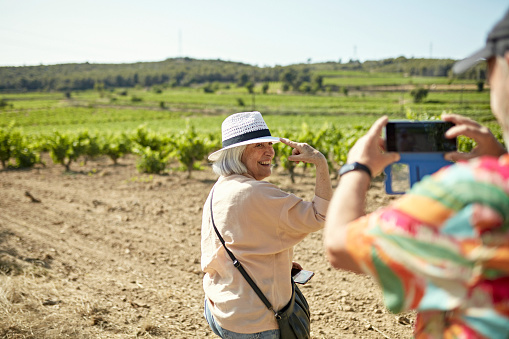 Waist-up view of smiling woman in hat and casual attire smiling at man capturing memories with smart phone as they walk through vineyard.