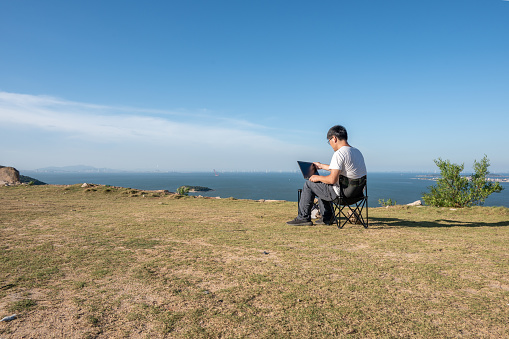 An Asian man uses a computer on a simple desk and chair in the wild by the sea