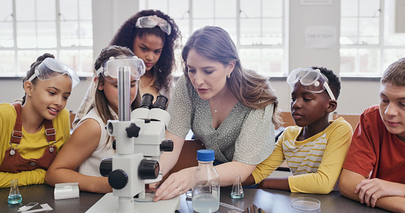 Teenage girl with classmates doing science experiment in laboratory. High school students examining liquid in test tubes. They are wearing protective eyewear and lab coats.
