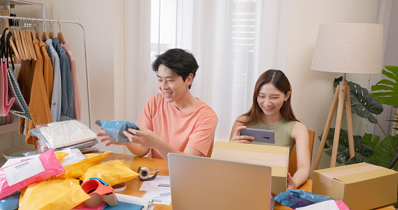 young asian couple small business owners packing product at home for online retail store order delivery to customer -  man sorts package and woman uses phone scan shipping ticket on plastic courier parcel bag and carton