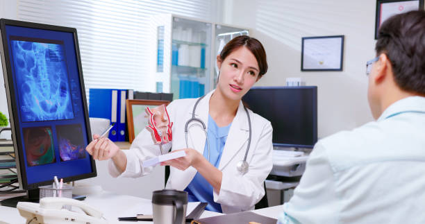 doctor explain rectum model attractive young brunette ponytail female asian doctor wear white coat and stethoscope explaining rectum model to sick male patient at clinic - an xray film display on computer anus stock pictures, royalty-free photos & images
