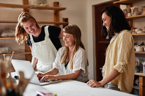 istock Pottery store owner talking and laughing, joking and having fun with her team and is happy due to business progress. Group of women laughing and smiling in a clay shop while typing on a laptop 1413756708