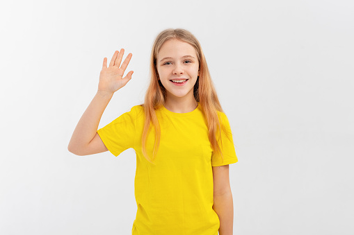 A friendly teen girl says hello, waves hand and smiles happy at you, stands in empty yellow tshirt over white background