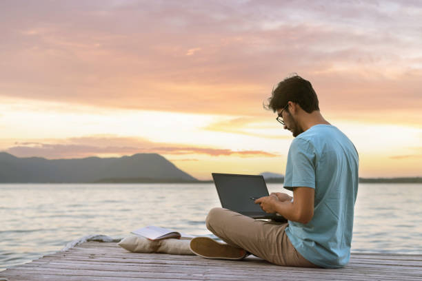 young digital nomad man sitting on wooden pier - on beach laptop working imagens e fotografias de stock