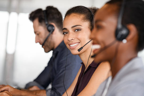 Customer service woman smiling at call center Portrait of smiling customer service operator with headset sitting with colleagues and attending calls in office. Beautiful black woman telemarketing agent working in call center with team and looking at camera. Smiling african american customer service representative sitting in a row. call center stock pictures, royalty-free photos & images