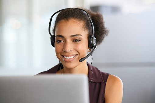 Call center agent with headset working on support hotline in modern office. Young black businesswoman in conversation with customer during video call. African american business woman working remotely on laptop and videocall.
