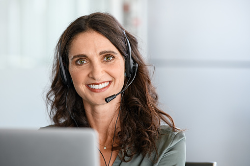 Call center agent with headset working on support hotline in modern office. Happy customer care representative in conversation with clients over headset looking at camera. Portrait of woman consulting clients on video call using laptop from home.