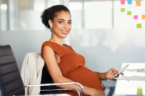 femme d’affaires noire enceinte utilisant un téléphone au bureau pendant qu’elle travaille - anticipation smiling touching image technique photos et images de collection