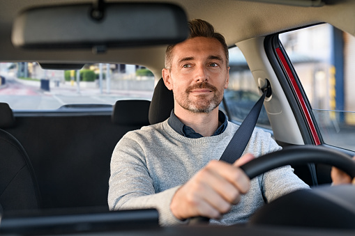 Mid adult casual man smiling while driving car and looking at mirror for reverse. Happy man feeling comfortable sitting on driver seat in his new car. Smiling mature businessman with seat belt on driving vehicle for transport and copy space.