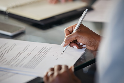 Close up of hand of black business woman signing agreement document in modern office. African american businesswoman signing contract. Woman in formal clothing sealing paperwork after deal and successful negotiations.