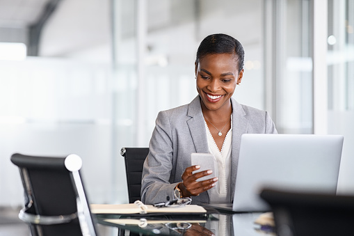 African american businesswoman using smartphone while working on laptop in office. Smiling mature black business woman checking phone while working. Successful woman entrepreneur browsing on phone in office.