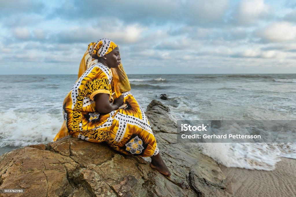 African woman with long dress sitting on stone cliff African woman with long dress sitting on stone cliff after the coast in Accra Ghana West Africa Ghana Stock Photo