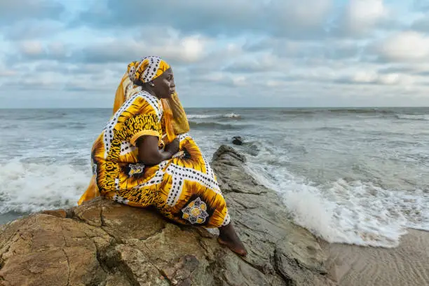 African woman with long dress sitting on stone cliff after the coast in Accra Ghana West Africa