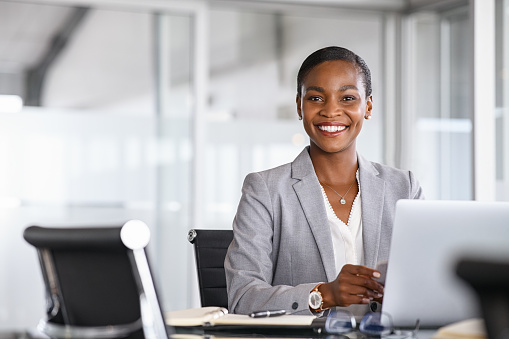 Portrait of a cheerful businesswoman sitting at desk in modern office and looking at camera. Attractive and successful black business woman sitting at conference table with laptop. Smiling african american executive using laptop while working from office with copy space.