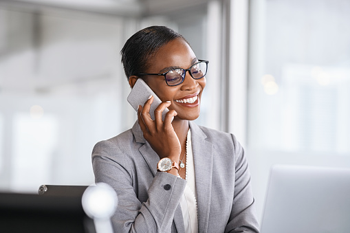 Mid adult businesswoman talking on cellphone in office. Smiling african american business woman in happy conversation on mobile phone. Mature confident black woman entrepreneur sitting at table and working from office.