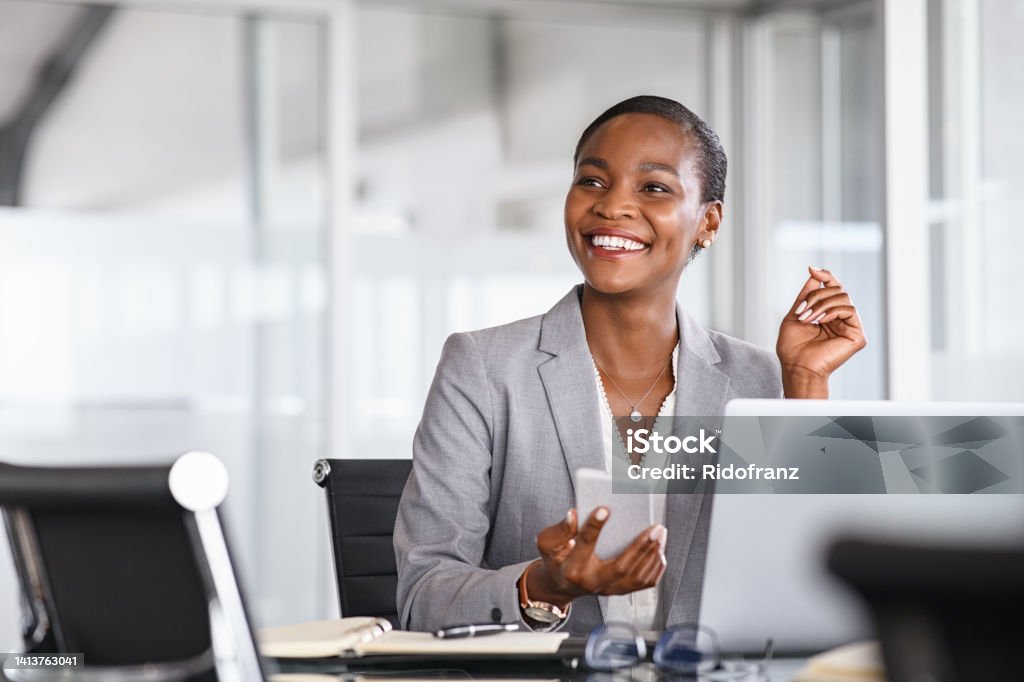 Smiling businesswoman looking up while working African black business woman using smartphone while working on laptop at office. Smiling mature african american businesswoman lookingup at copy space while working on phone. Successful woman entrepreneur think about new business ideas. Business Stock Photo