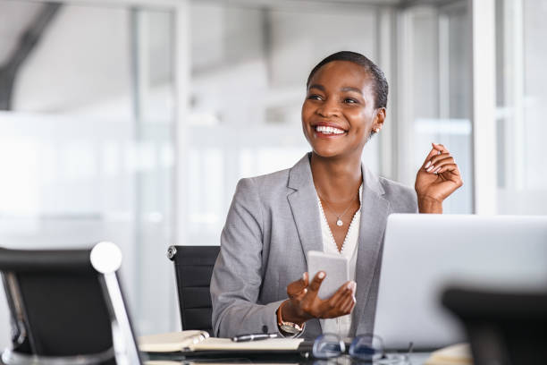 Smiling businesswoman looking up while working African black business woman using smartphone while working on laptop at office. Smiling mature african american businesswoman lookingup at copy space while working on phone. Successful woman entrepreneur think about new business ideas. ceo stock pictures, royalty-free photos & images