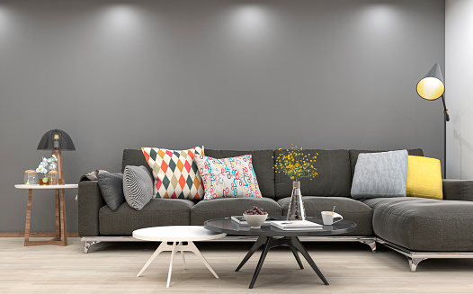 Elegant living room with gray textile sofa, two tables, decoration (multi-colored pillows, vase with flowers fruit, books) on hardwood parquet floor in front of empty medium gray plaster wall with copy space. An elegant lamp in the corner, a table lamp on a small coffee table on the other side of the sofa and reflectors that illuminate tha wall. Slight vintage effect added. 3D rendered image.