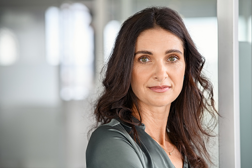 Portrait of hispanic successful woman entrepreneur smiling. Close up face of confident businesswoman standing in modern office with copy space. Mature satisfied formal woman looking at camera while smirking.