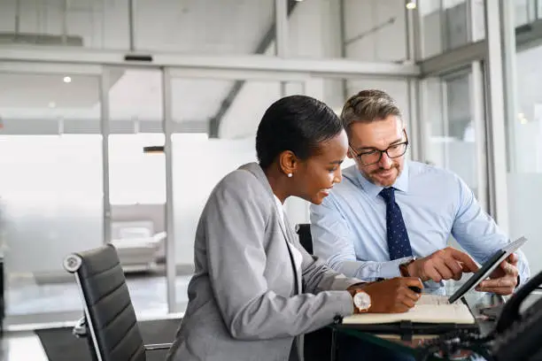 Mature businessman discussing work with black manager writing notes in diary. Confident business man in formal clothing working with african american woman showing data on digital tablet. Smiling professional business people working together on new business strategy.