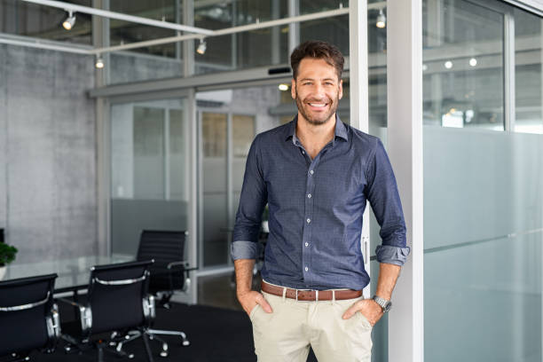 Successful business man looking at camera in office Portrait of mature successful man with hands in pocket standing outside office conference room. Handsome businessman standing in office while looking at camera. Young satisfied man in formalwear standing in boardroom with copy space. mid adult men stock pictures, royalty-free photos & images