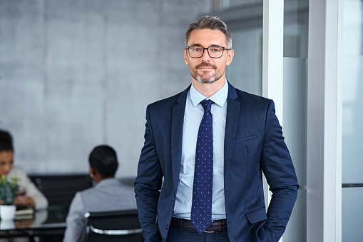 Portrait of handsome mid adult business man standing in modern office. Successful mature entrepreneur in formal clothing looking at camera with satisfaction. Confident man in suit with eyeglasses and beard standing with hands in pocket and looking at camera in office.