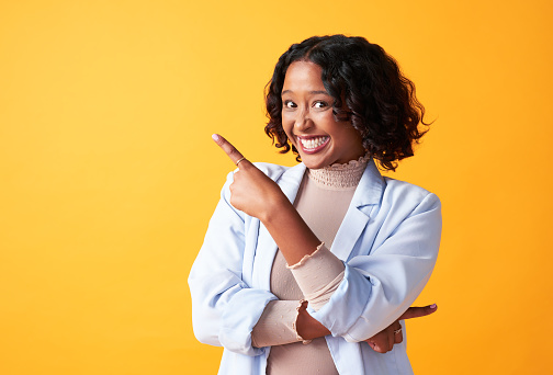 Excited, stylish and trendy woman pointing her finger, showing direction or raising her hand standing against a yellow background. Trendy, happy and cheerful female making hand gesture with copyspace