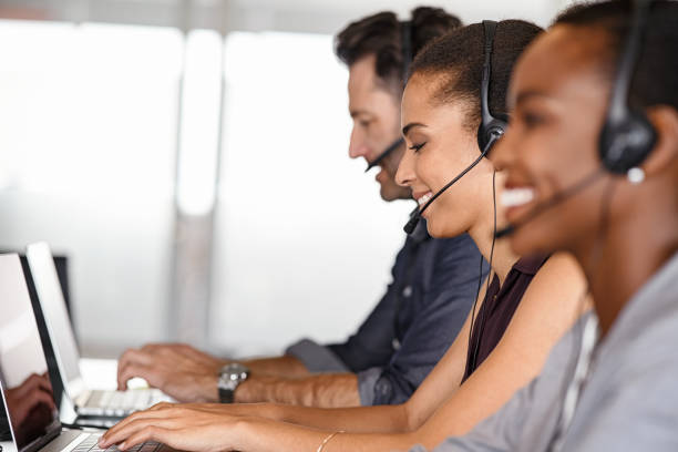 Customer service agents on call Smiling multiethnic people with headsets using computer and smiling while working in office. Young man and woman operators talking on headset with clients. Group of telemarketing customer service team working while sitting in a row. call centre stock pictures, royalty-free photos & images