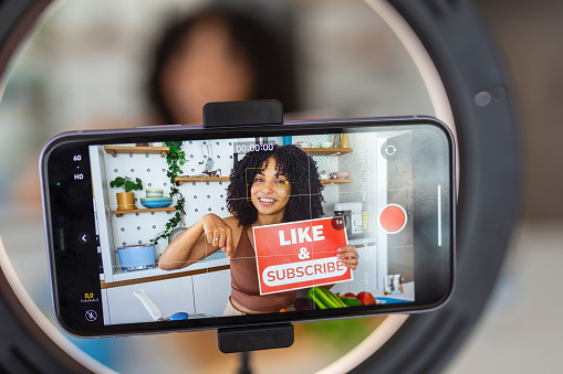 An Afro-Caribbean woman showing a creative recipe with healthy fresh ingredients from her kitchen at home, using her smart phone to film the cookery demonstration