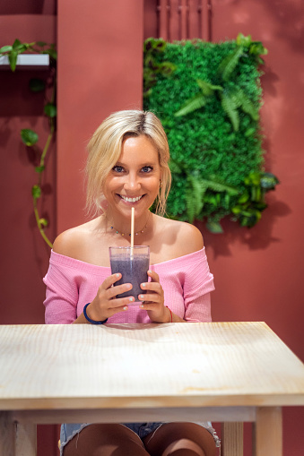 Happy woman smiling at camera while drinking a delicious smoothie in a cafe. Healthy lifestyle concept.