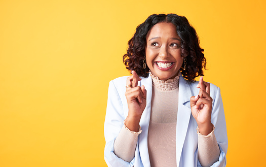 Happy black woman keeping fingers crossed and hoping for the best against a yellow background. Excited African American female making a wish while waiting for good news, a promotion or loan approval