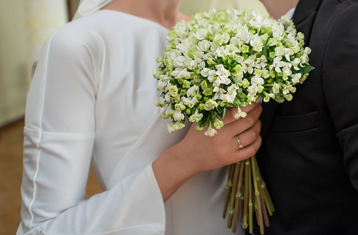 Close view of bridegroom in suit, bride in white dress hugging him with bouquet of flowers. Close view of wedding ring