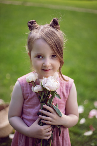 portrait of a beautiful baby girl in a pink dress standing with a bouquet of flowers pions on a green meadow in summer.