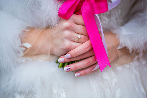 The bride holds a bouquet with pink ribbons in her hands. White dress and wedding ring