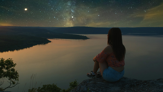 The girl sitting on the mountain top above scenic river on starry sky background