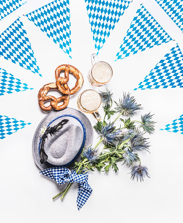 Beer Fest layout with traditional Bavarian white blue fabric,  pretzel , cups of draft beer, Bavarian hat, party flag garland, decoration and bunch of alpine flowers on white background. Top view.