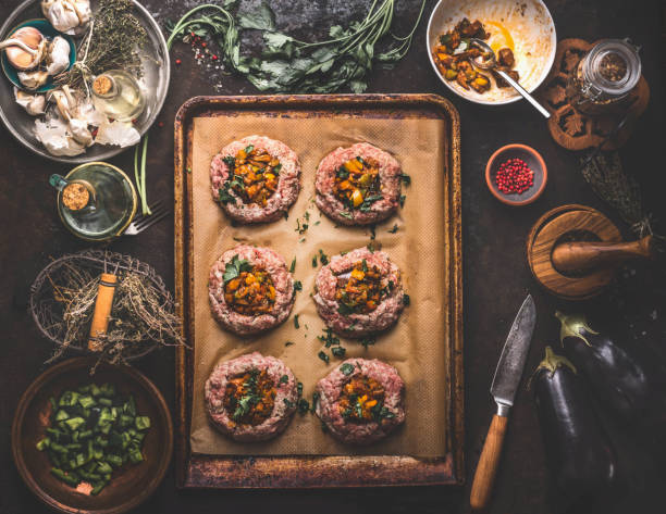 Preparation of minced meat patties in bagel shape filled with roasted vegetables on baking sheet on rustic kitchen table with herbs, spices and utensils. stock photo