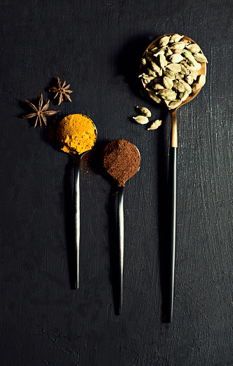 Various spices, flavors and herbs lined up on a black table for cooking a tasty meal. Ingredients for a flavorful dish on spoons with a rustic background. Assortment of Indian seasoning
