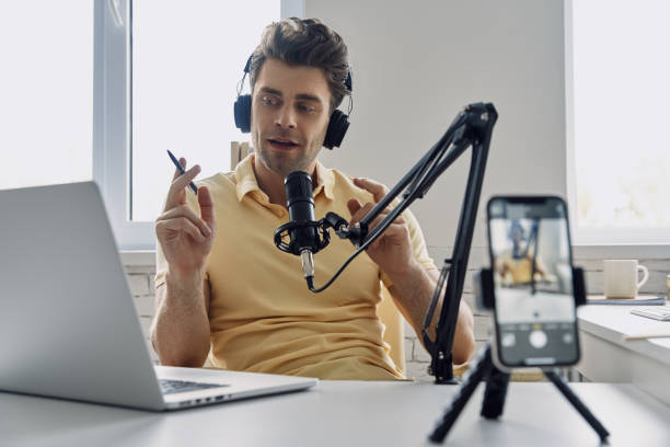 istock Young man in headphones using microphone and looking at laptop while recording podcast in studio 1413800069