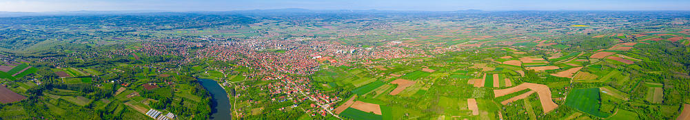 Panoramic above view, overhead, on lake, forest, colorful hilly landscape with cottage settlement, several greenhouses on cultivated arable plots, farmland, city in the distance.
