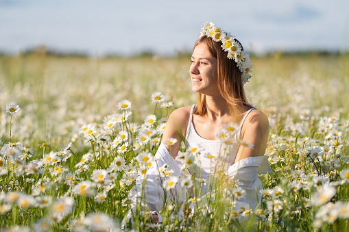 A beautiful girl is enjoying life in a daisy field at sunset. A young woman with a wreath of daisies on her head smiles happily in nature.