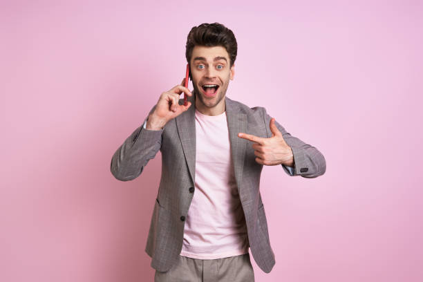 istock Excited man in suit talking on mobile phone and pointing it while standing against pink background 1413802654