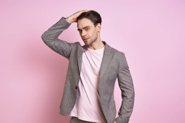 Handsome young man in suit holding hand in hair while standing against pink background stock photo