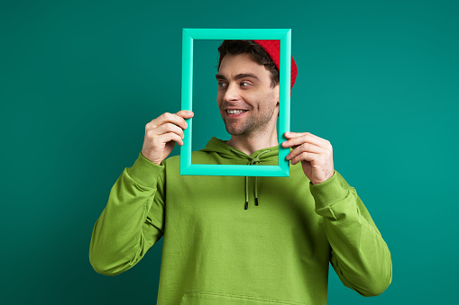 Playful young man looking through a picture frame while standing against green background